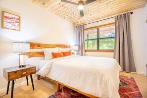Ein Schlafzimmer mit Holz-Kopfteil, einem Bett mit weißer Bettwäsche, einem Nachttisch mit Lampe, einem Fenster mit Vorhängen und Blick auf Grünflächen.
