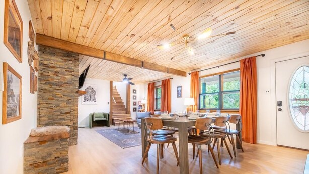 Una habitación amplia con techo de madera, una mesa de comedor con sillas, una escalera y un gran ventanal con cortinas.