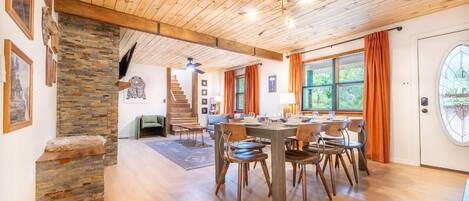 Une pièce spacieuse avec un plafond en bois, une table à manger avec des chaises, un escalier et une grande fenêtre avec des rideaux.