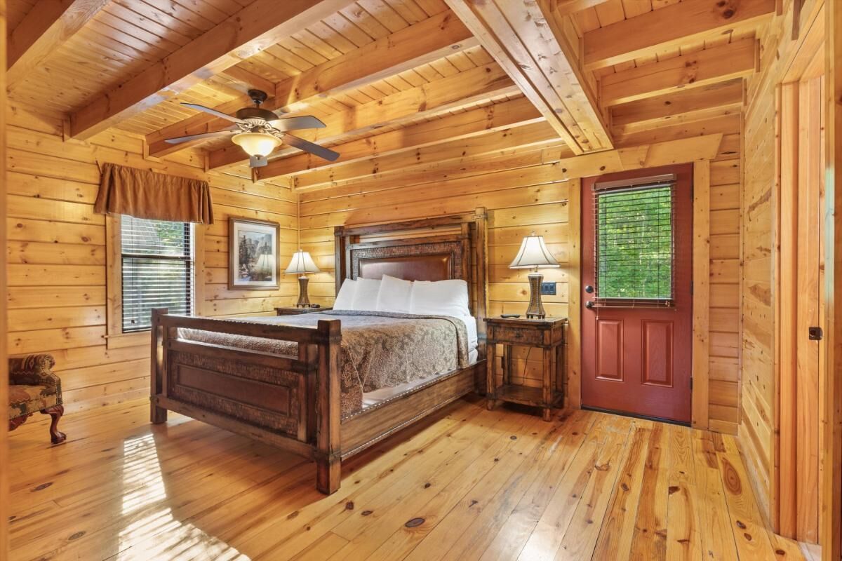 A wooden cabin bedroom with a bed, two bedside tables, a ceiling fan, and a door with a window.