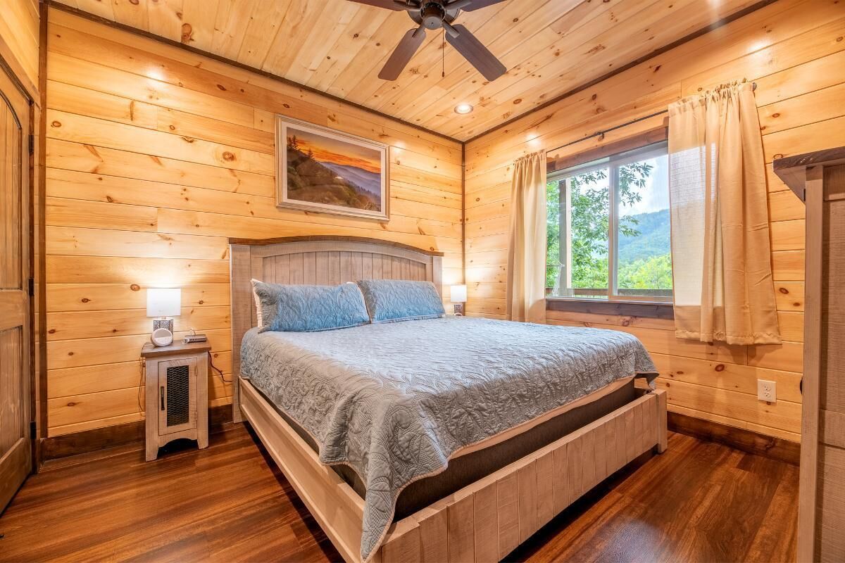 A wooden cabin bedroom with a bed, a nightstand, a ceiling fan, and a window with curtains.