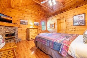 A wooden cabin interior with a stone fireplace, a bed with a colorful blanket, a dresser, and framed pictures on the walls. - Z - Southern Style (Sevierville)