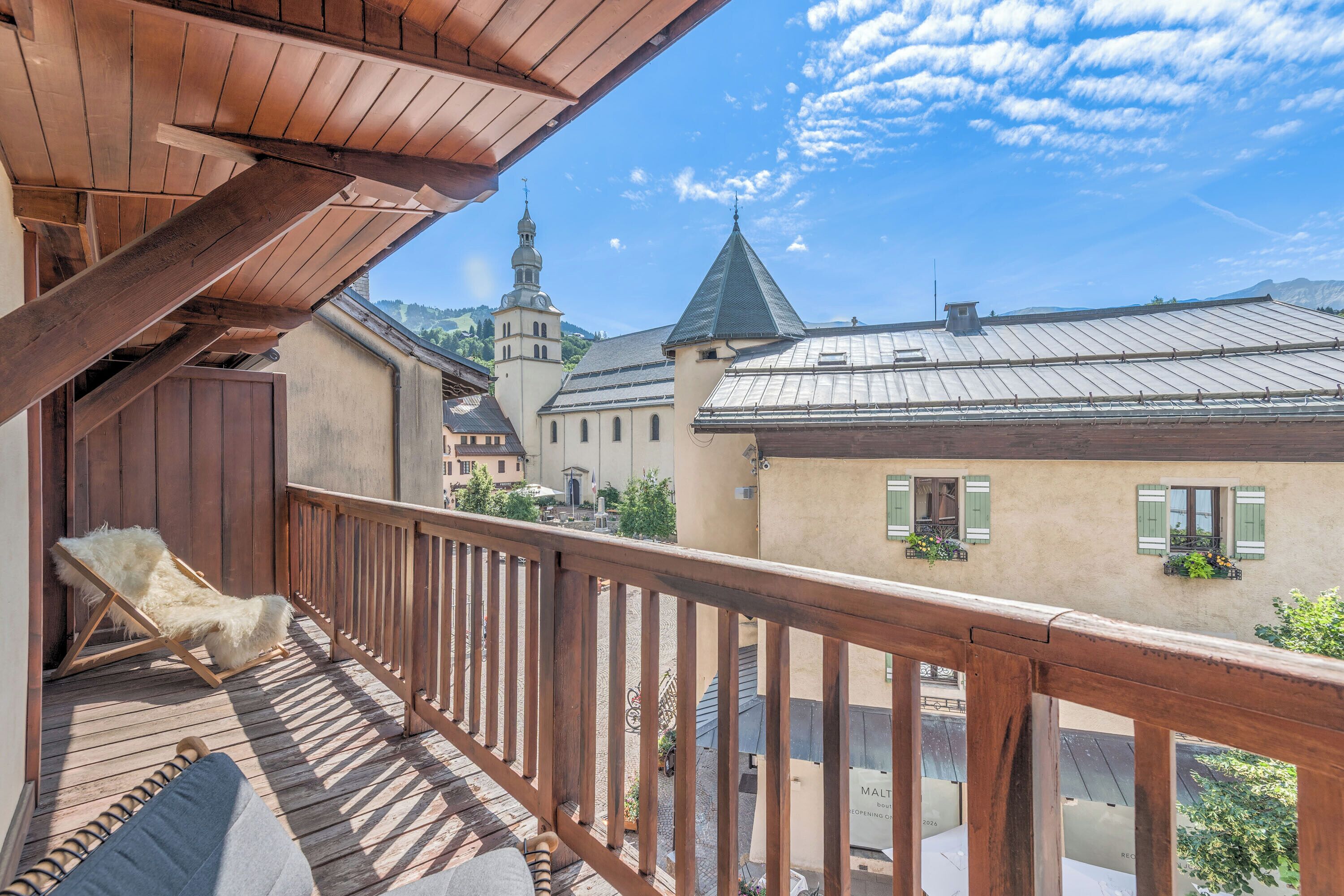 A balcony with wooden railings, a white furry chair, and a view of a town with buildings and a church tower.
