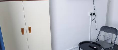 Una habitación con un armario blanco, una mesita redonda con un control remoto, una silla gris y un televisor montado en la pared.