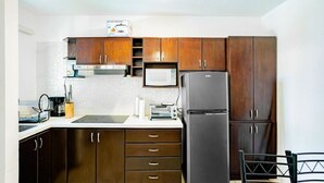 A compact kitchen with wooden cabinets, a microwave, a refrigerator, and a small dining area with a chair. - Cocomarindo 66 - Stylishly Renovated 1BR (Guanacaste Province)