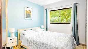 A bedroom with a bed, a nightstand, a lamp, a framed picture on the wall, and a window with curtains. - Cocomarindo 66 - Stylishly Renovated 1BR (Guanacaste Province)