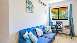A blue sofa with patterned cushions, a desk with a chair, and a window with curtains. - Cocomarindo 66 - Stylishly Renovated 1BR (Guanacaste Province)