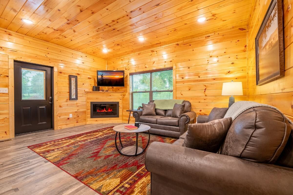 A log cabin interior with a fireplace, leather sofa, TV, and a large window.