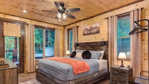 A wooden cabin bedroom with a bed, a ceiling fan, a window with curtains, and a painting on the wall. - Dreamcatcher Lodge (Sevierville)