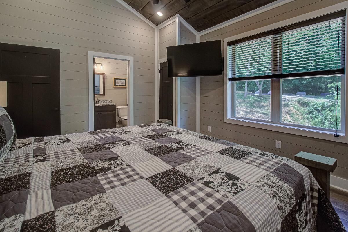 A bedroom with a large bed, a television mounted on the wall, a window with blinds, and a door leading to another room.