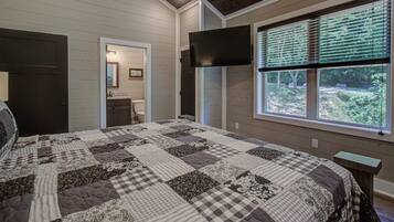 A bedroom with a large bed, a television mounted on the wall, a window with blinds, and a door leading to another room.