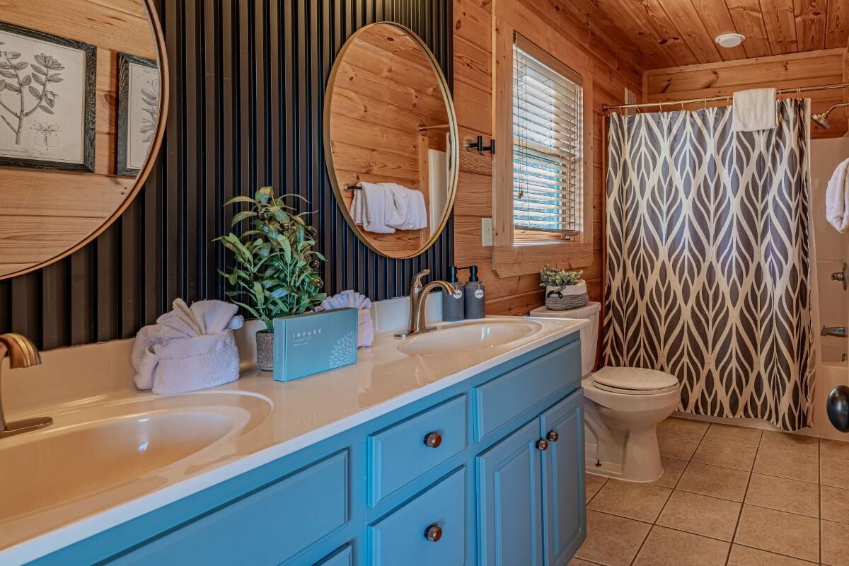 A bathroom with wooden paneling, a double sink vanity, a toilet, and a shower curtain with a leaf pattern.