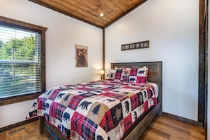 A bedroom with a wooden ceiling, a bed with a bear-themed quilt, a window with blinds, and a 'DO NOT FEED THE BEARS' sign. - Black Bear-y Ridge (Sevierville)