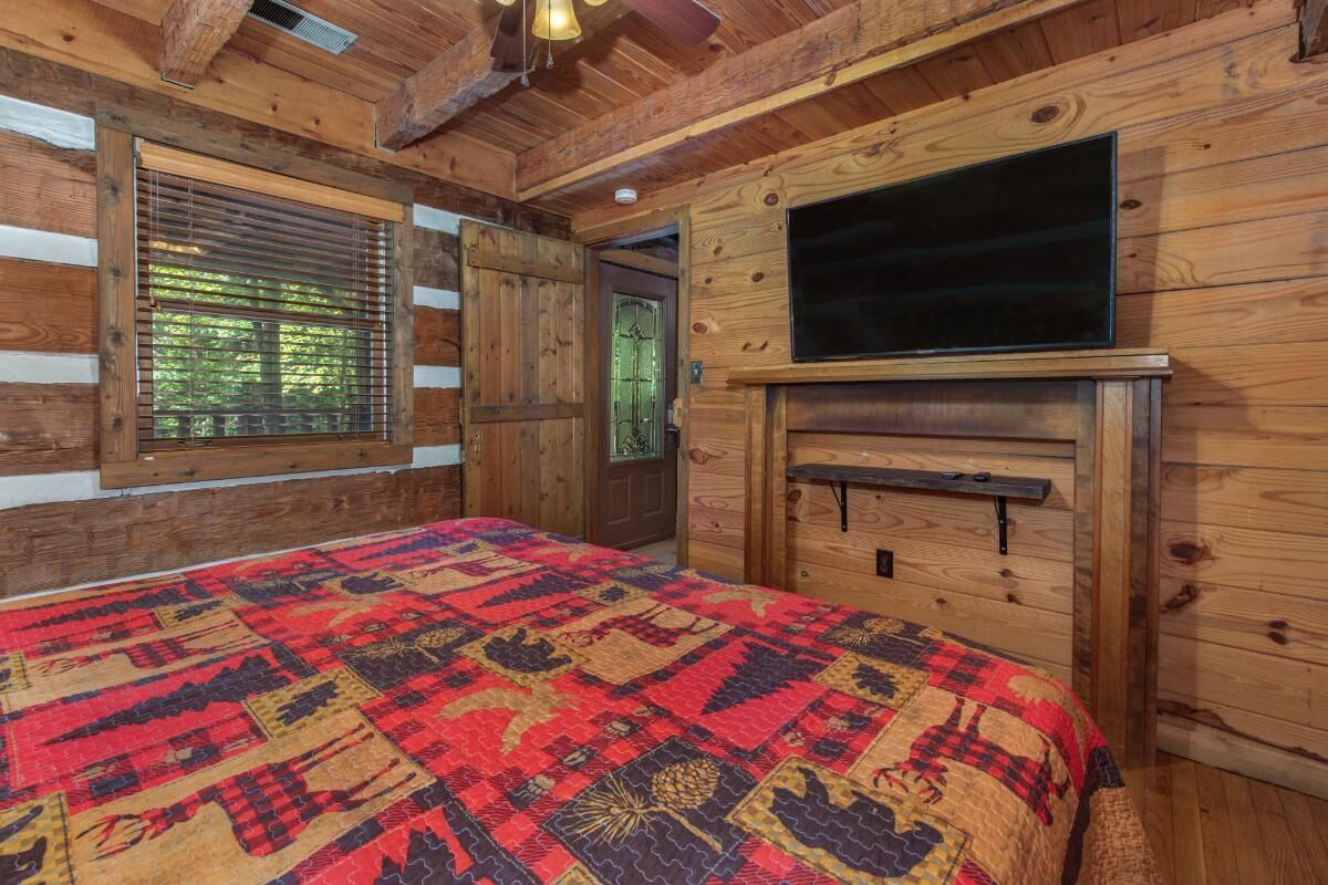 Ein Schlafzimmer in einer Blockhütte mit einem großen Bett, einem an der Wand befestigten Fernseher und einem Fenster mit Jalousien.