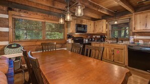 A rustic cabin interior with a wooden dining table, chairs, a kitchen with a microwave, and a large window with blinds. - Whistling Woods Hideaway (Sevierville)