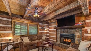 A log cabin interior with a stone fireplace, a ceiling fan, and a television. - Whistling Woods Hideaway (Sevierville)