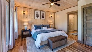 Quarto com teto de madeira, uma cama com um banco cinzento, duas luminárias de cabeceira e arte de parede com pinhas.