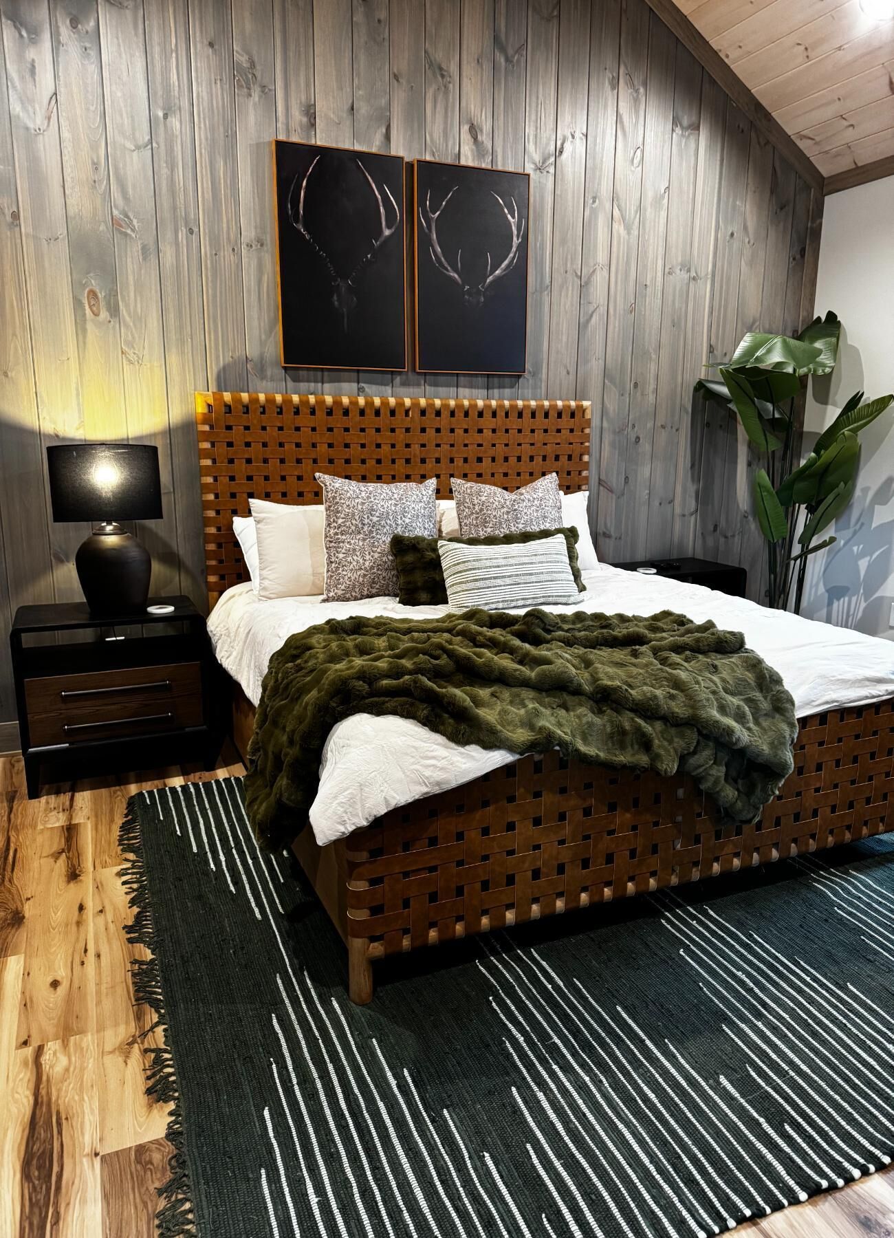 A bed with a woven headboard, a bedside table with a lamp, and a large plant in a room with wooden walls.