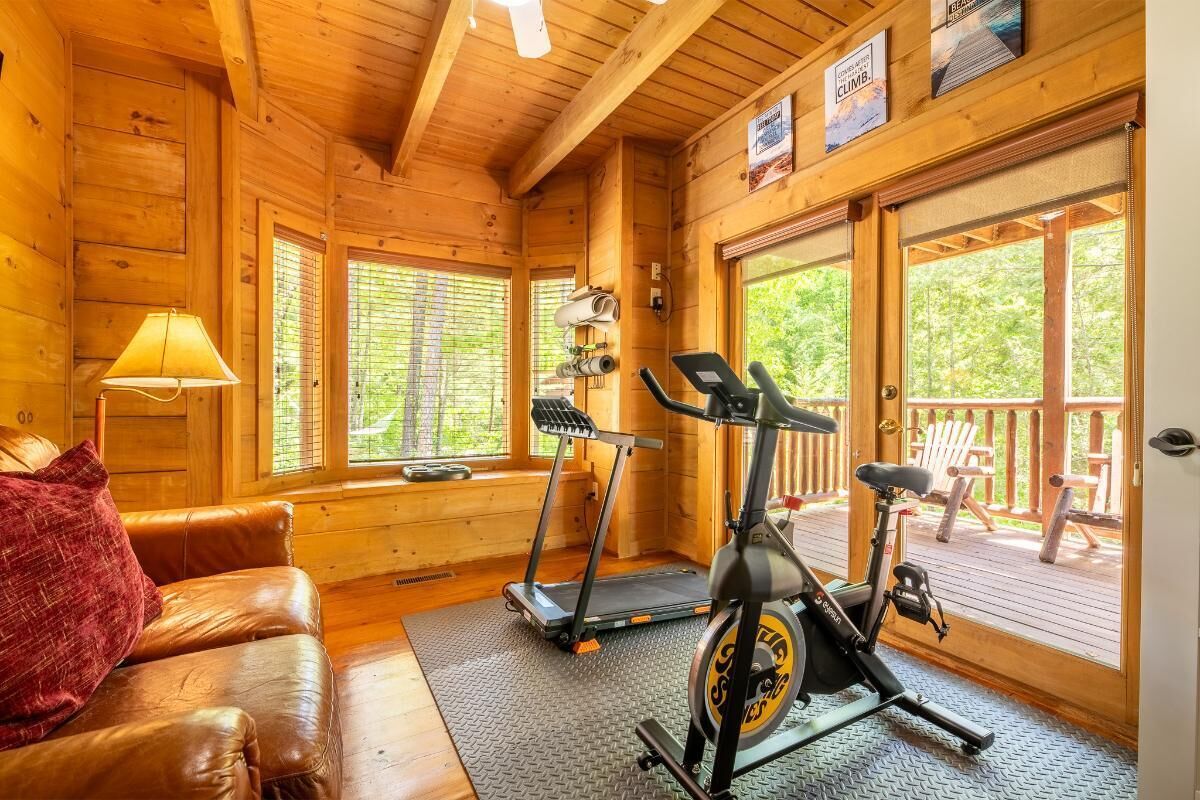 A wooden cabin interior with a treadmill, exercise bike, and a couch with a red pillow.