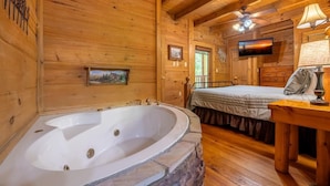 Ein Holzblock-Innenraum mit einer Steine Badewanne, einem Bett, einem Fernseher und einem Deckenventilator.