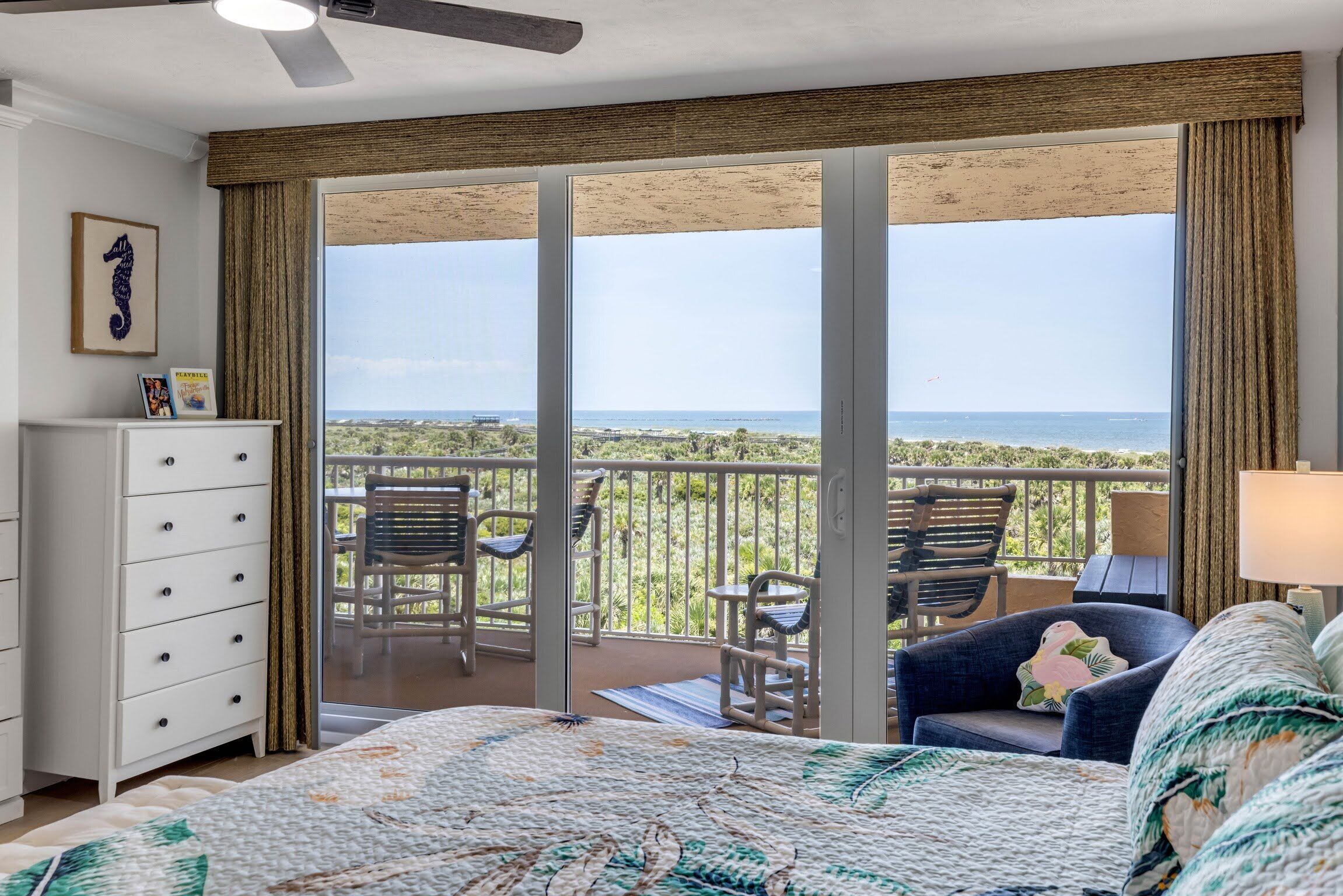 Une chambre dotée d’un grand lit, d’une commode, d’un ventilateur de plafond et d’un balcon avec des chaises et une vue sur l’océan.