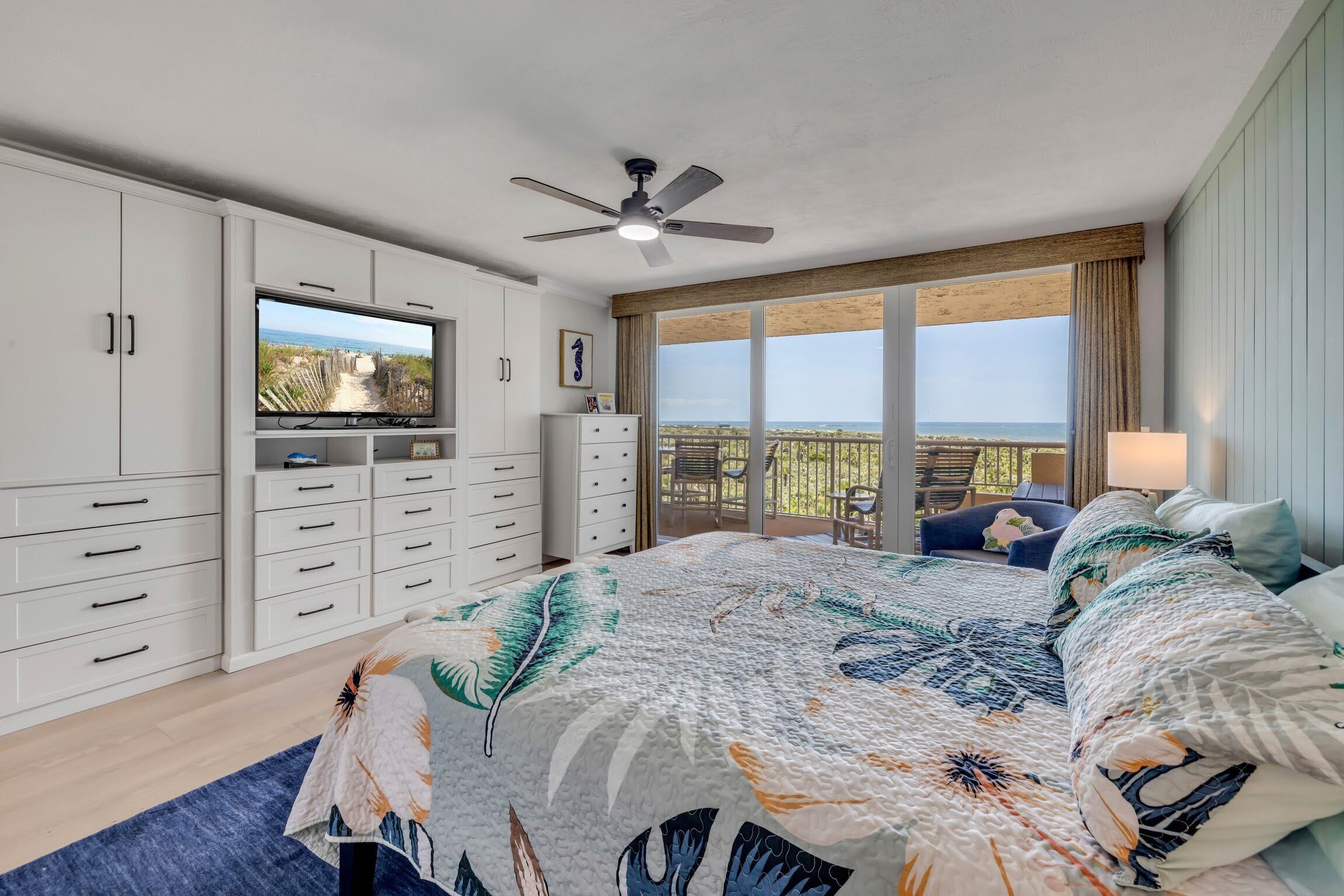 A bedroom with a large bed, a TV, a ceiling fan, and a balcony with a view.