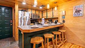 A kitchen with wooden cabinets, a stainless steel refrigerator, a microwave, and a coffee machine. There are bar stools and a sign that says 'DON'T FEED THE BEARS'. - Z - Black Bear Hideout (Gatlinburg)