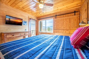 Ein Zimmer in einer Holzhütte mit einem Bett, einem Fenster, einem Deckenventilator und einem Wandfernseher.