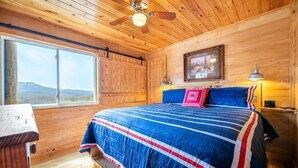 A wooden cabin bedroom with a bed, a ceiling fan, a window with a mountain view, and a framed picture on the wall. - Z - Black Bear Hideout (Gatlinburg)
