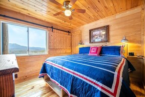 Ein Schlafzimmer einer Holzhütte mit einem Bett, einem Deckenventilator, einem Fenster mit Blick auf Berge und einem gerahmten Bild an der Wand.