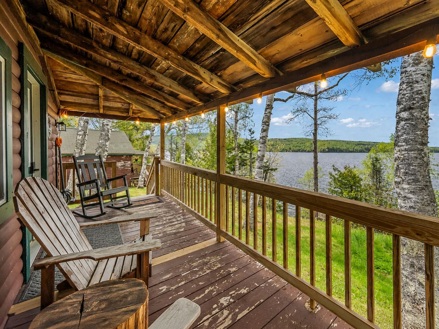 A wooden deck with chairs and a table, overlooking a lake with birch trees.