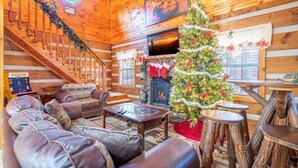 A log cabin interior with a fireplace, Christmas tree, leather sofa, and a staircase. - Antlers Haven (Pigeon Forge)