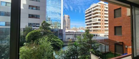 A view from a window with a balcony, overlooking a cityscape with modern buildings and a clear sky.