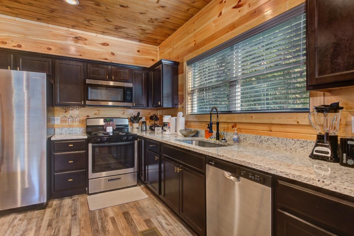 A modern kitchen with dark cabinetry, stainless steel appliances, and a large window with blinds.