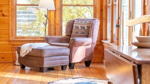 Interno di una baita in legno con una poltrona in pelle, un poggiapiedi, una lampada e vista sugli alberi attraverso ampie finestre.