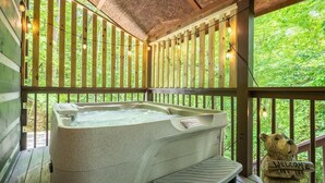 Un jacuzzi en una terraza de madera con un letrero de un oso que dice 'Bienvenido', rodeado de barandas de madera y luces colgantes.