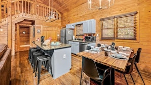 Interior de una cabaña de madera con comedor, cocina y una escalera que sube a un nivel superior.