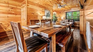 A wooden interior with a dining table set for four, chairs, a ceiling fan, and a window with blinds. - Mountain Getaway (Pigeon Forge)