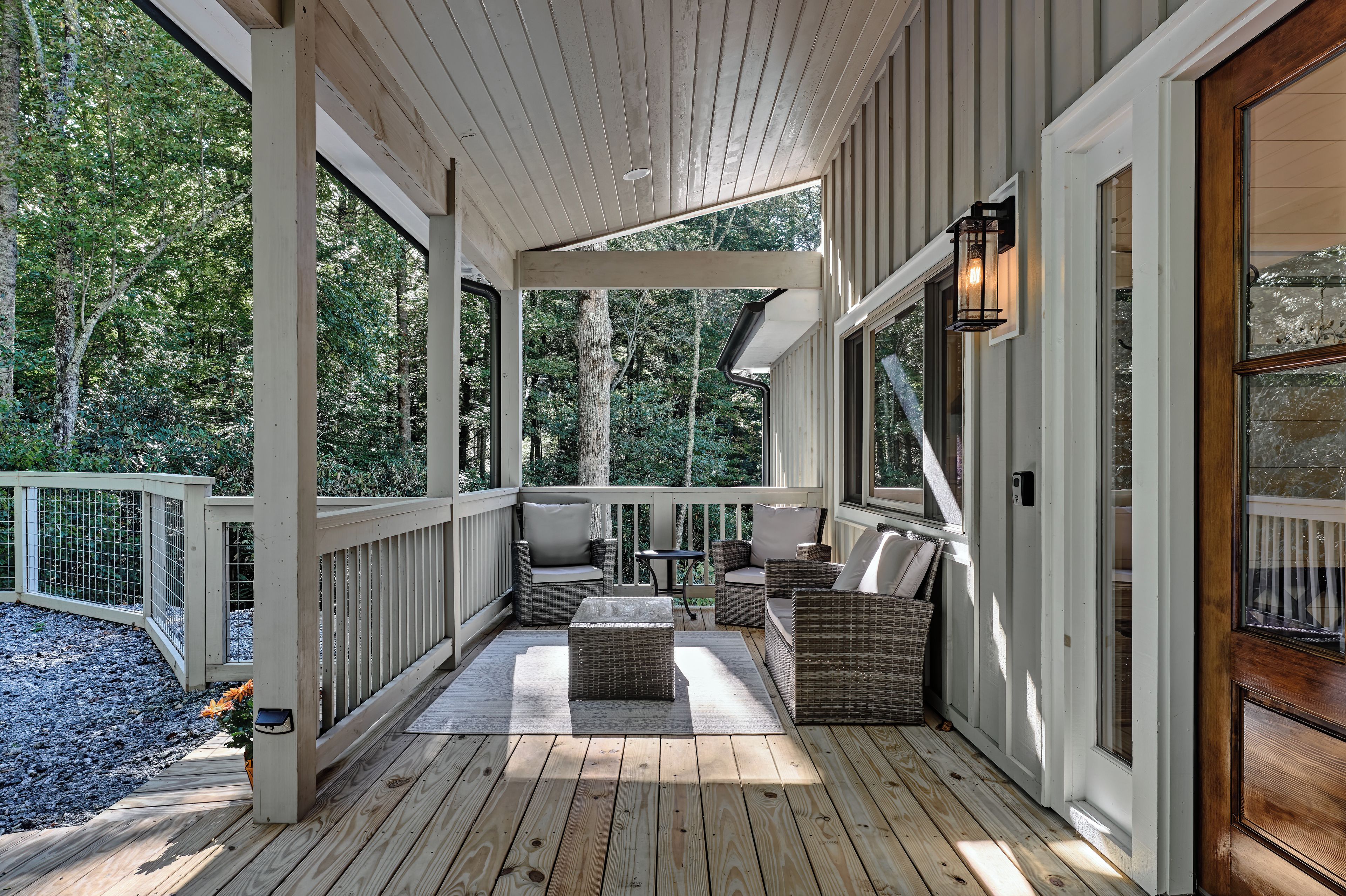 A covered porch with wooden flooring, wicker furniture, and a view of a forested area.