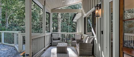 A covered porch with wooden flooring, wicker furniture, and a view of a forested area.