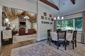 A spacious living area with a fireplace, a dining table set for four, and a large window offering a view of the outdoors. - Private Pond, Mountain Escape (Highlands)