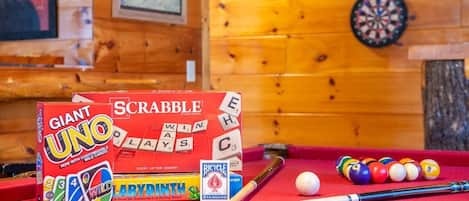A pool table with billiard balls, a cue stick, and two game boxes: Scrabble and Giant UNO.