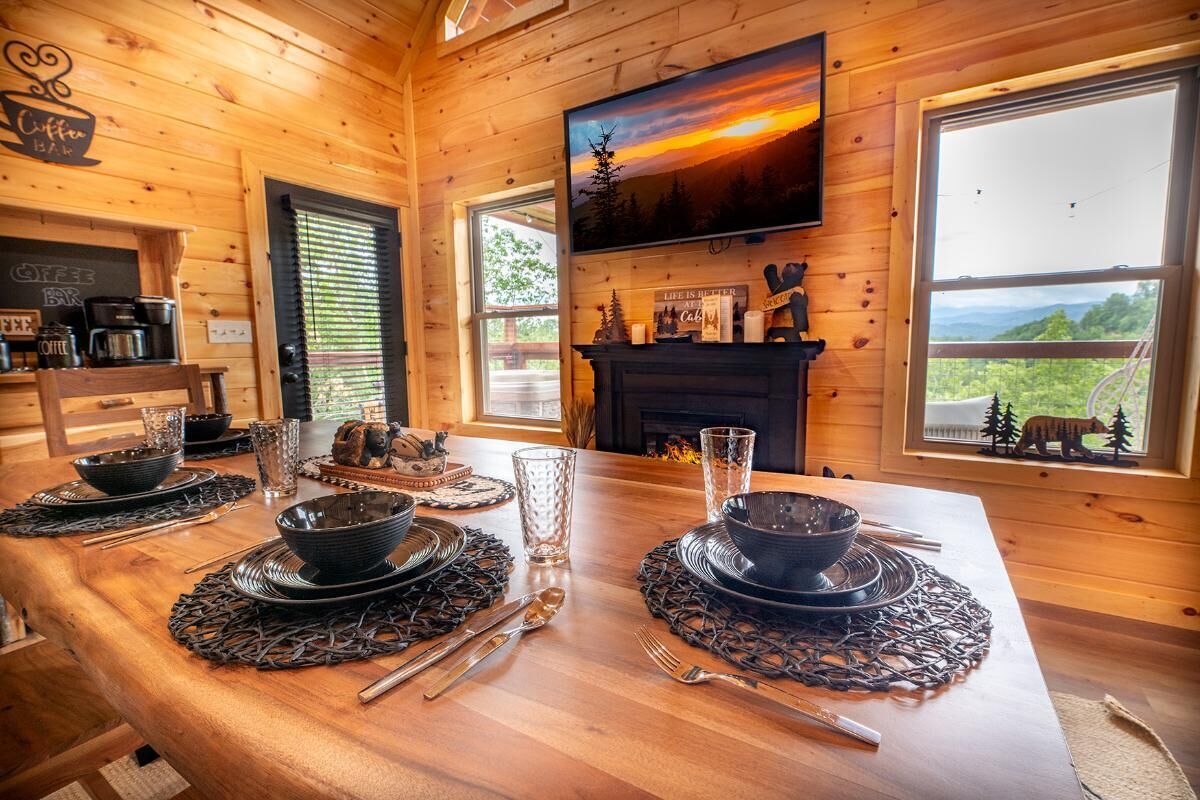 A wooden interior with a dining table set for four, a fireplace, a TV mounted on the wall, and a scenic view through the window.