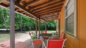 Een overdekt terras buiten, met een tafel, stoelen, een hangplant en een raam met luiken.