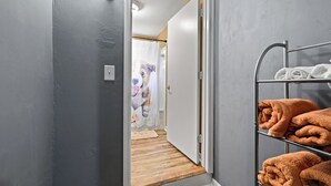 Een gang met een metalen handdoekrek met opgevouwen handdoeken, een deur naar een kamer en een lichtschakelaar aan de muur.