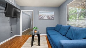 Een woonkamer met een blauwe bank, een klein tafeltje met een plant, een wandtelevisie en een schilderij aan de muur.