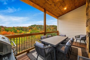 A covered outdoor patio with a table and chairs, a grill, and a view of autumn foliage. - Lookout Lodge (Sevierville)