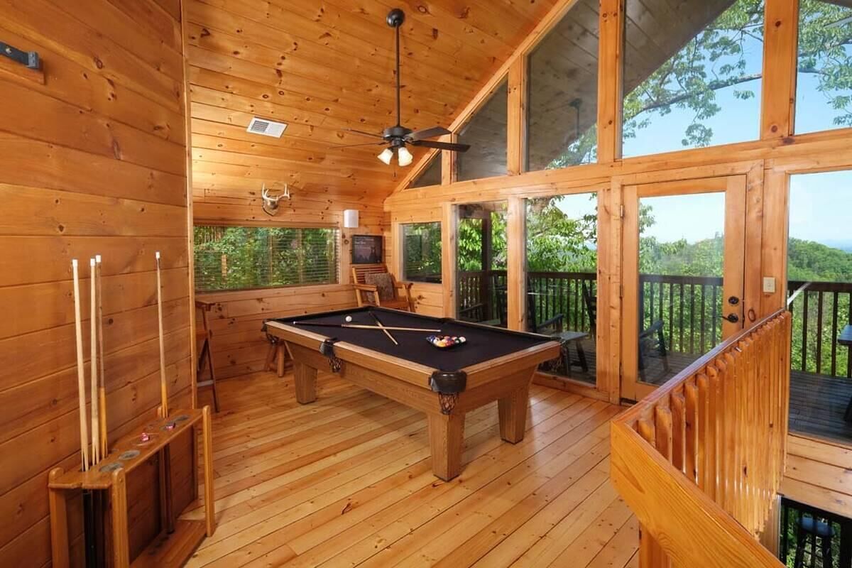 A wooden interior with a pool table, ceiling fan, and large windows overlooking a deck.