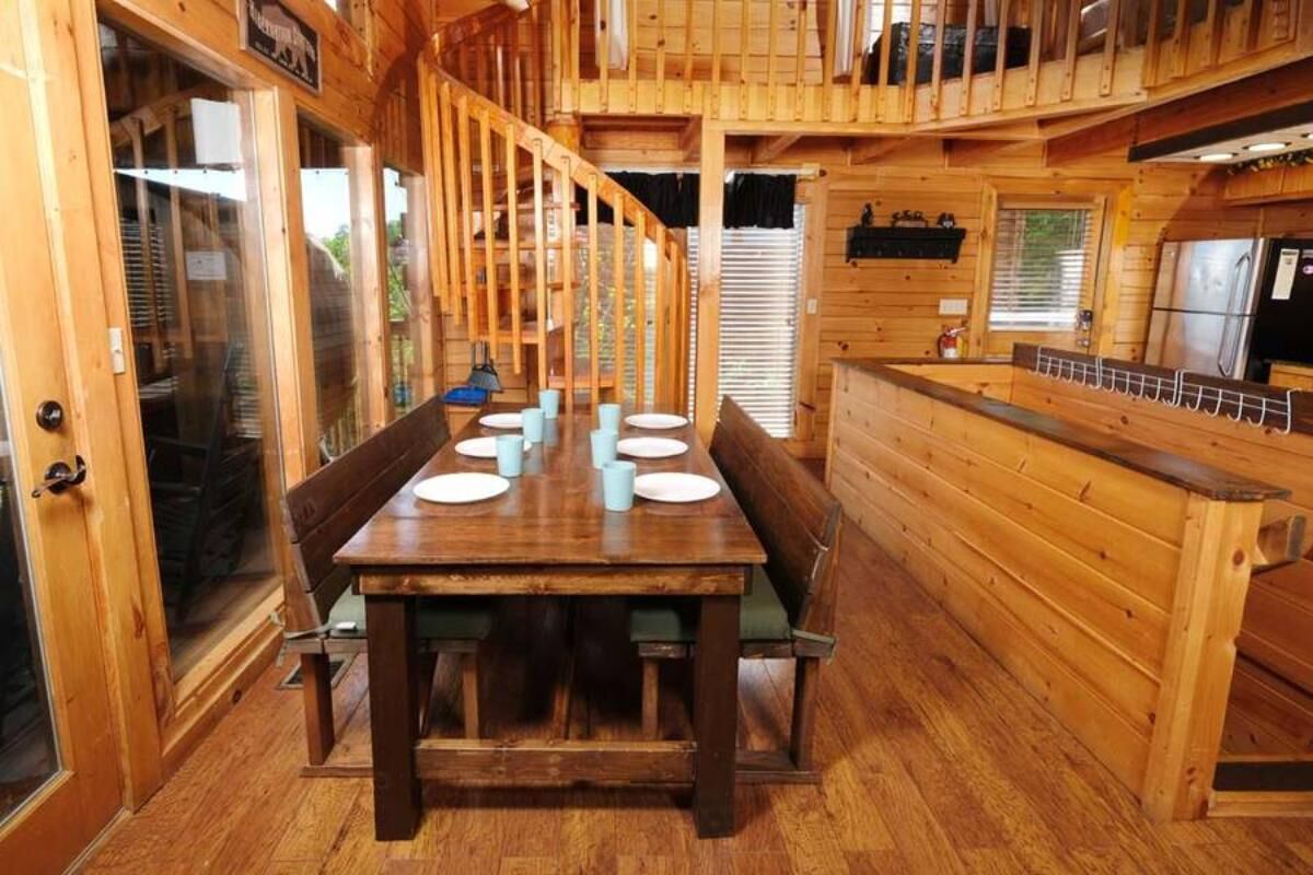 A wooden cabin interior with a dining table set for six, a kitchen area, and a staircase.