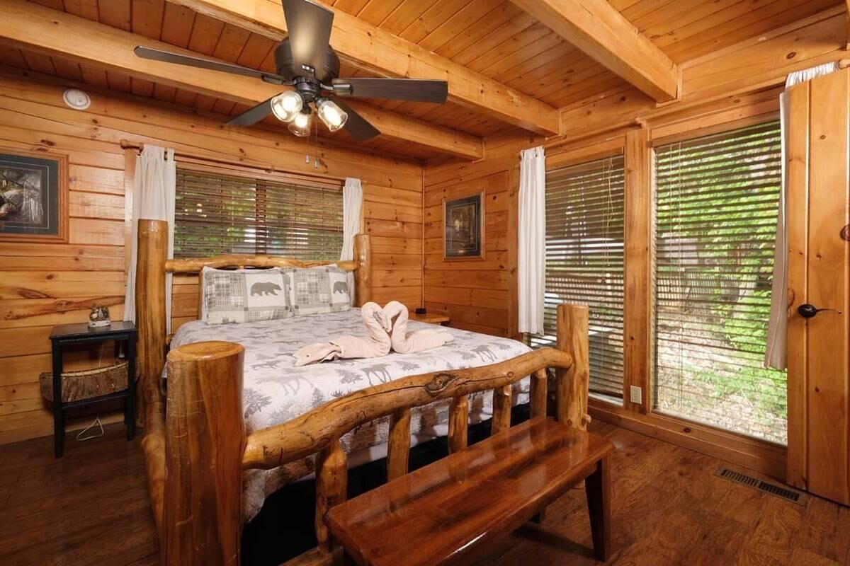 A wooden cabin bedroom with a bed, a bench, a ceiling fan, and large windows.